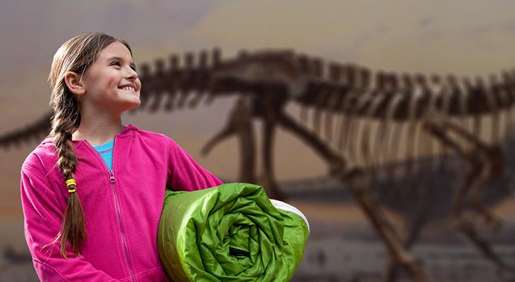 A child holds a sleeping bag in front of a dinosaur skeleton.