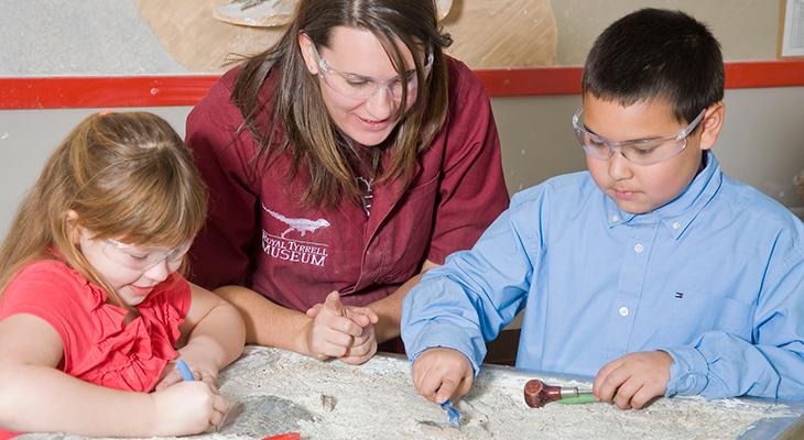 An educator supervises two children uncovering fossils. 